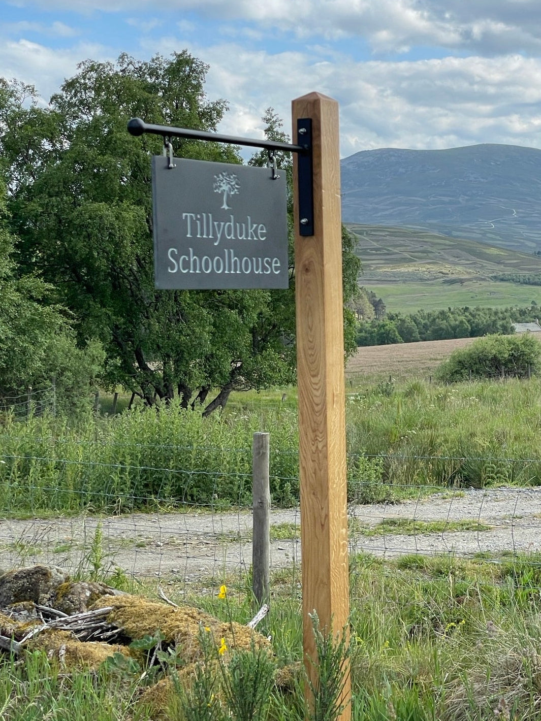 Corian hanging sign with engraved text in a natural setting with mountains and sky.